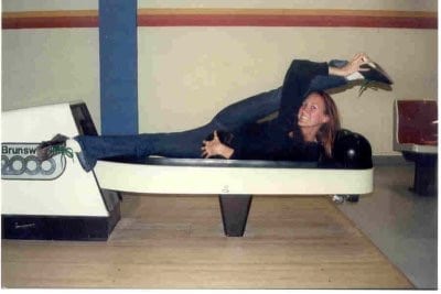 same white woman laying on bowling ball return machine, holding her right leg up into a split position (heel stretch.)