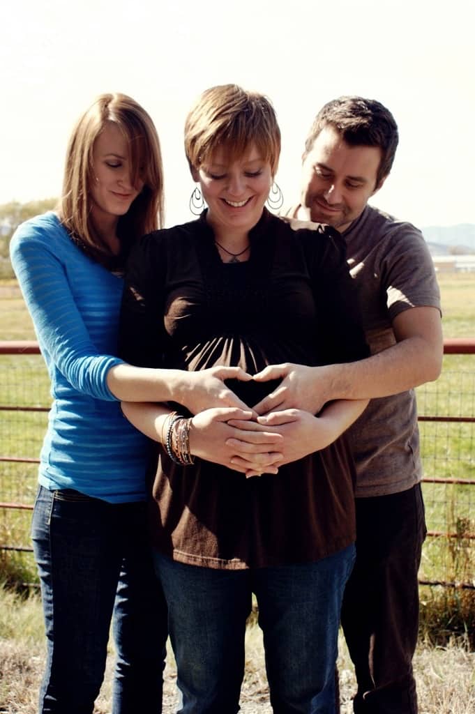Man and woman standing behind and at each side of a young pregnant woman. Man and woman's hands come together in a heart shape on pregnant woman's belly. Her hands are clasped together overlapping their hands. All are looking down at belly.