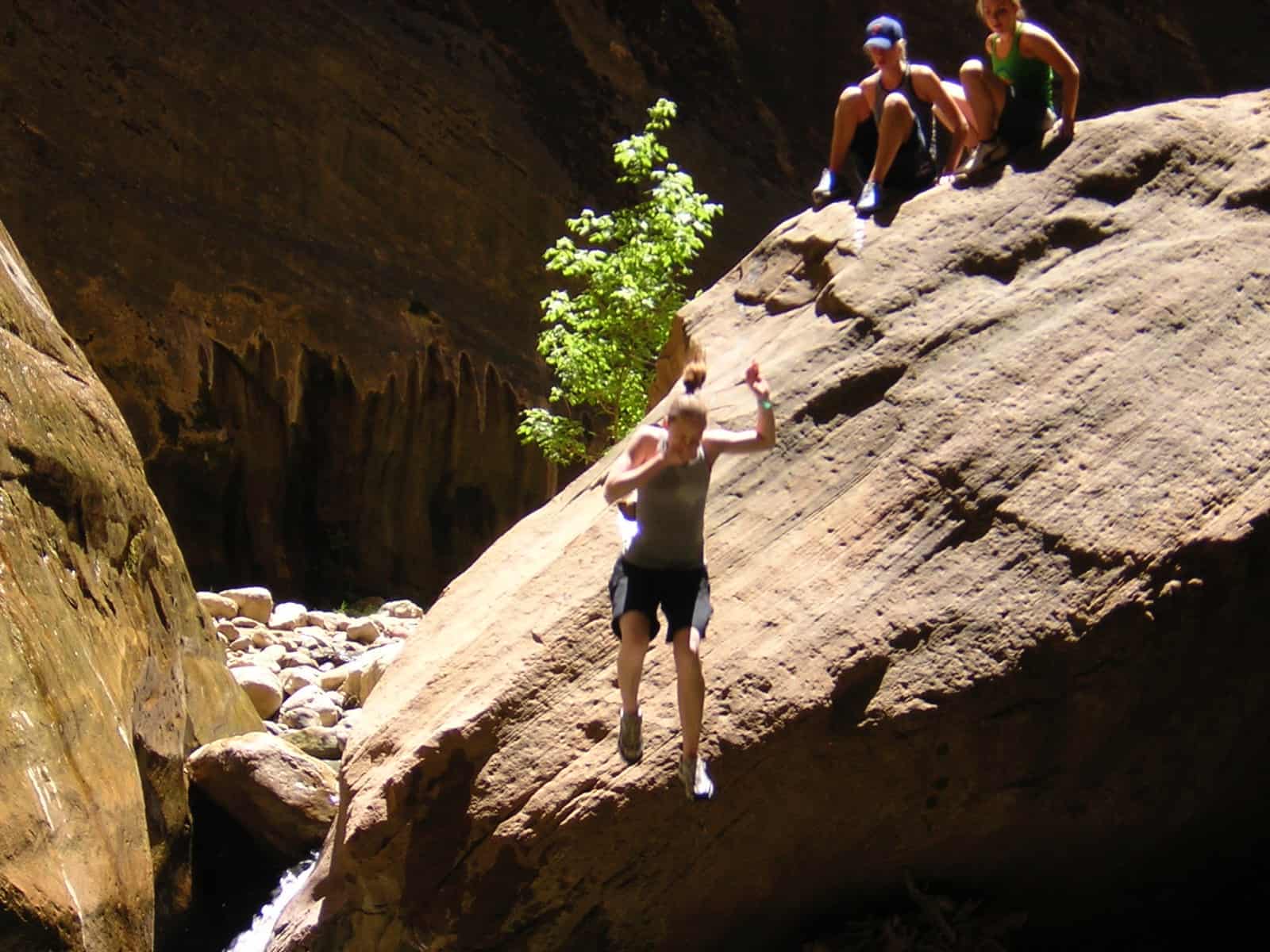 Two young women sitting on top of a boulder watching a third woman jump down holding her nose.