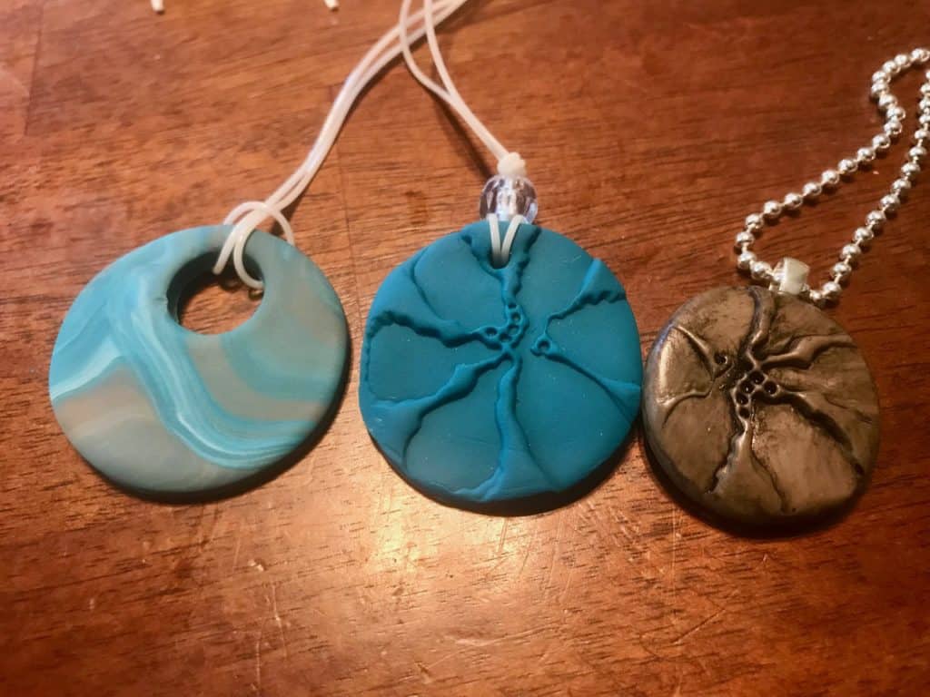 3 different clay pendents left to right: a blue, white, and grey marbled, round clay pendent, a deep blue round pendent with a flower pattern pressed in, same pendent as the middle one but paint a grey metallic.