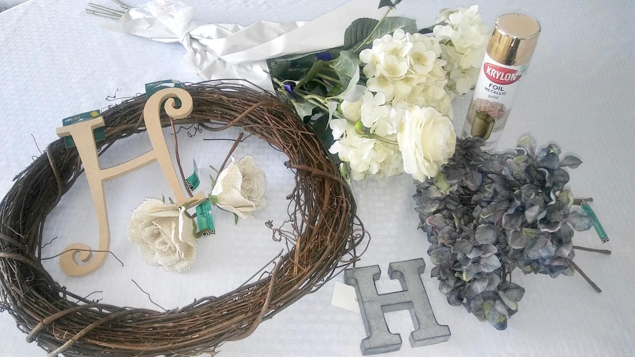 Craft supplies to make a wreath including a wicker wreath, a metal letter H, a cardboard letter H, spray paint in gold, florals in white and grey.