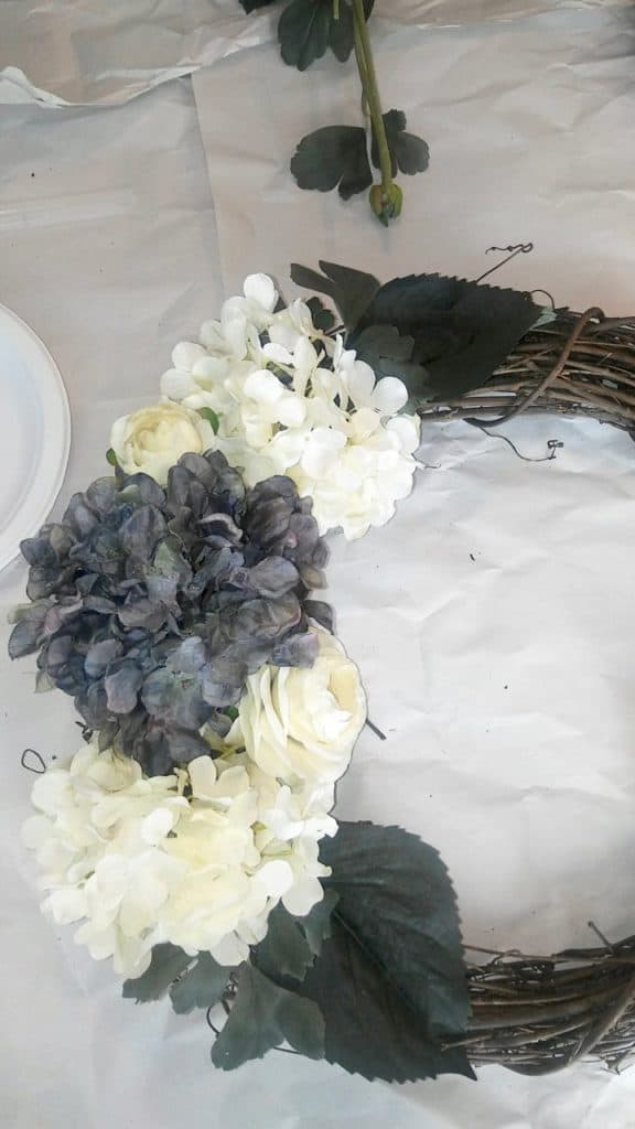 Small white flowers, full white roses, grey-ish hydrangeas, and a couple of leaves in varying sizes, sit on a wreath form. 