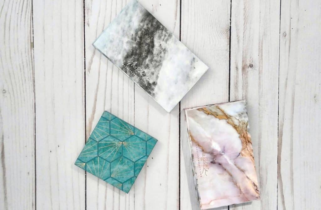 3 Seint beauty compacts with different looks. One with a black and white marble look. One with a pink, gold, white marble look. One in turquoise with a deeper turquoise hexagon pattern and gold lines.