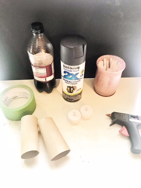 Toilet paper rolls, fake tea lights, hot glue gun and sticks, masking tape, black spray paint, and Dr. Pepper all sitting on a white table.