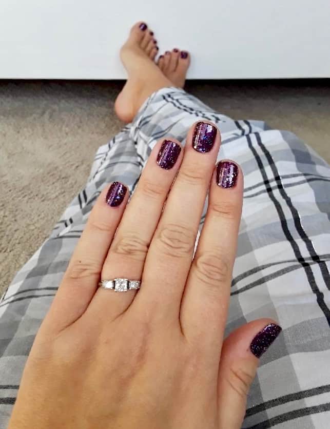 The focus of the photo is on a lady's hand wearing deep purple and glitter nail polish strips. She's sitting down with outstretched legs, wearing light gray plaid pj bottoms. You can see her bare feet in the background wearing the same nail polish.