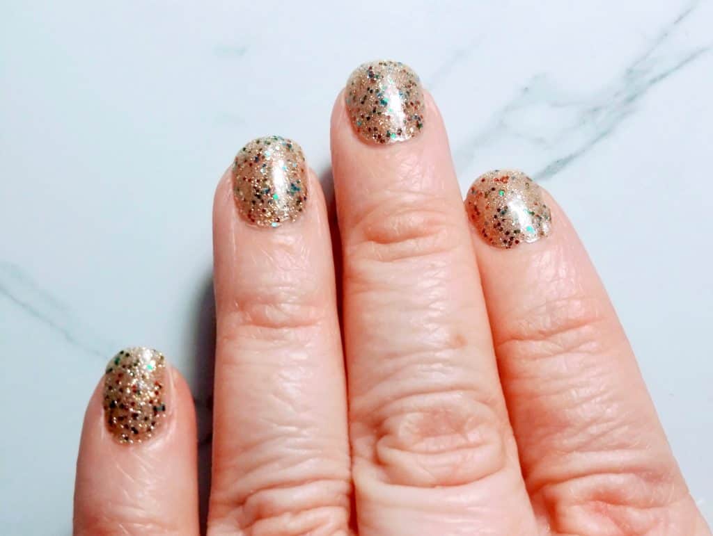 Four fingers with short nails, wearing gold and color glitter Color Street nail polish strips.