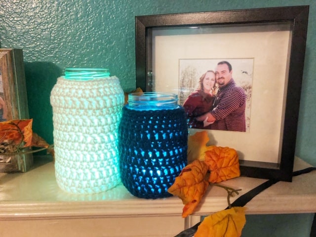 A large mason jar cover in a white crochet cozy sitting next to a blue crochet cozy on a smaller mason jar glowing from a candle inside the jars. 