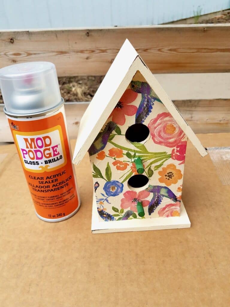 The finished decoupage and painted birdhouse outside and a can of Mod Podge brand clear acrylic sealer standing next to it.