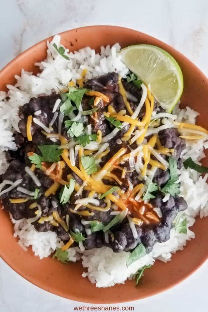 Burt orange bowl full of white rice topped with black beans, shredded cheese, chopped cilantro, a little red hot sauce with a lime wedge on the side sitting on a white counter top.