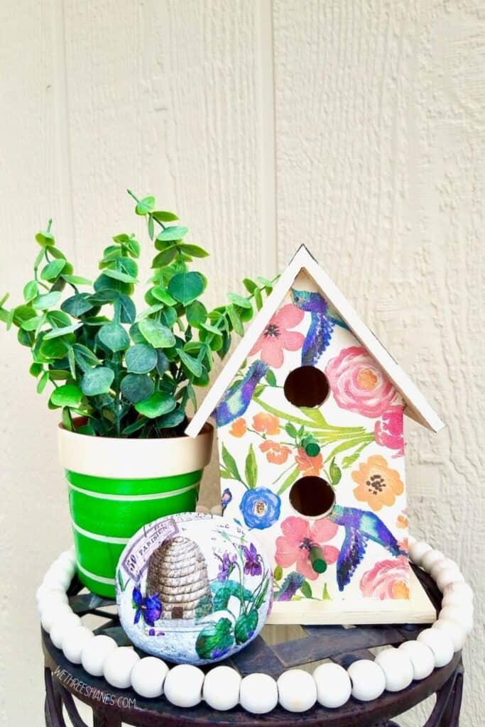 You can decoupage all kinds of things for spring and summer decor. A birdhouse decoupaged with flower napkins, a rock with a beehive and purple flowers, and a terra cotta pot with a green napkin with white stripes. | We Three Shanes