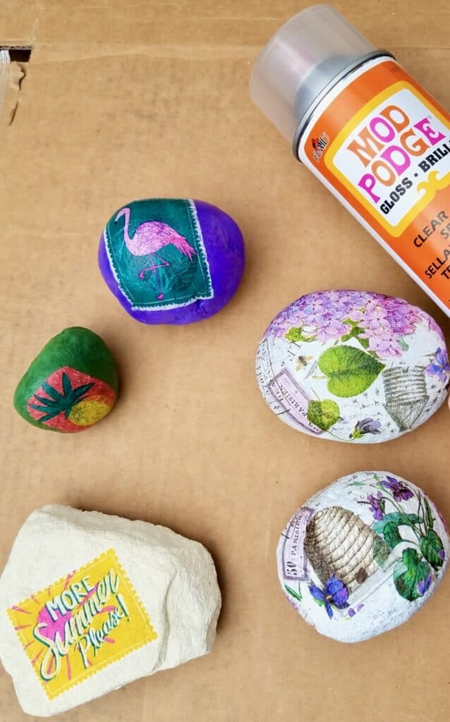 A purple rock with a pink flamingo, a green rock with a pineapple, two slightly bigger rocks with delicated purple flowers, leaves, and bee hives on them, and an even bigger white rock with the words "more summer please" decoupaged on and ready to be sealed.