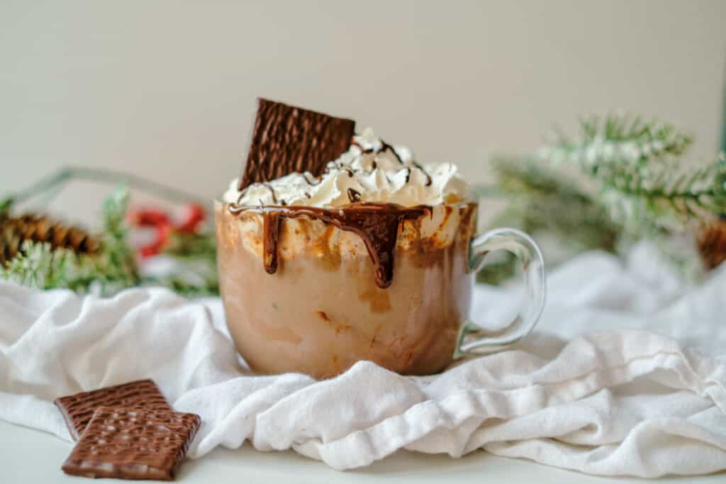 Clear mug of hot chocolate topped with whipped cream, drizzled with chocolate syrup, with an After Eight candy sticking out of the whipped cream.