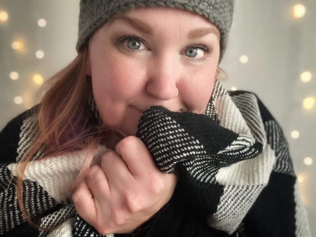 a woman with red hair, wrapped in a plaid blanket smiling.  She is wearing a grey beanie and there are some blurred out Christmas lights behind her.