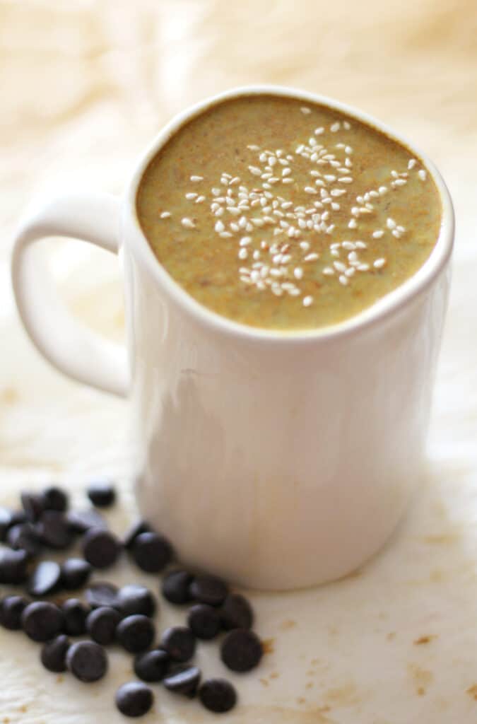 White mug full of yellow brown drink, topped with sesame seeds.