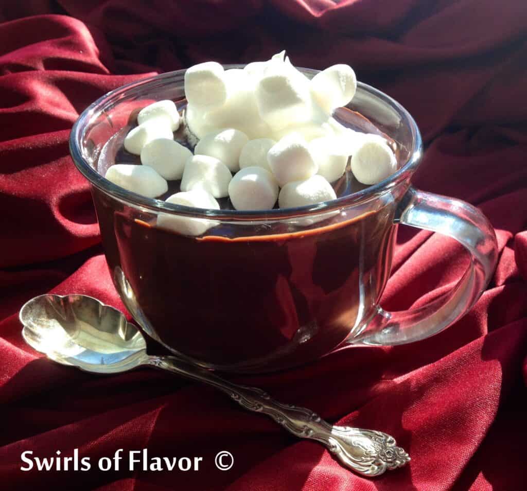 Clear mug of hot cocoa topped with mini marshmallows sitting on maroon velvet material.