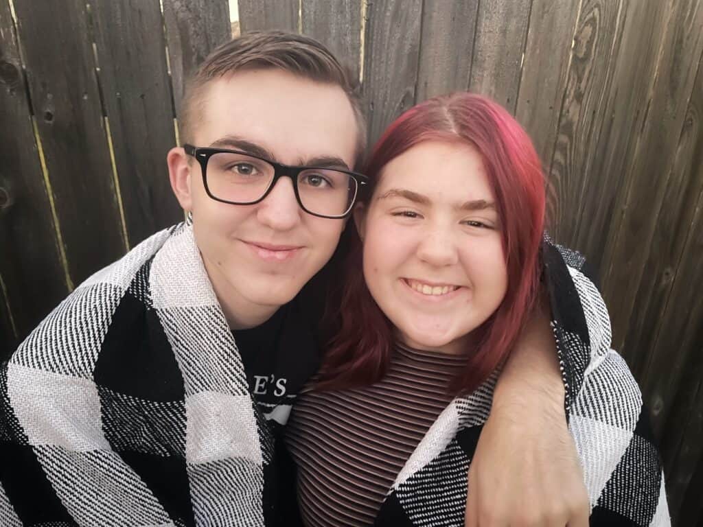 Two teenagers, a boy and a girl.  Boy on the left has his arm around the girl to the right and they have a blanket around them.