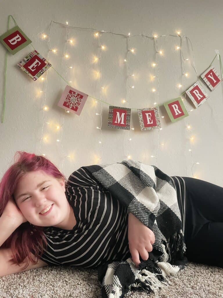 Christmas lights hung on a wall with a sign that says "Be Merry" across it.  A teenage girl with pink hair with a plaid blanket is laying on her side smiling.