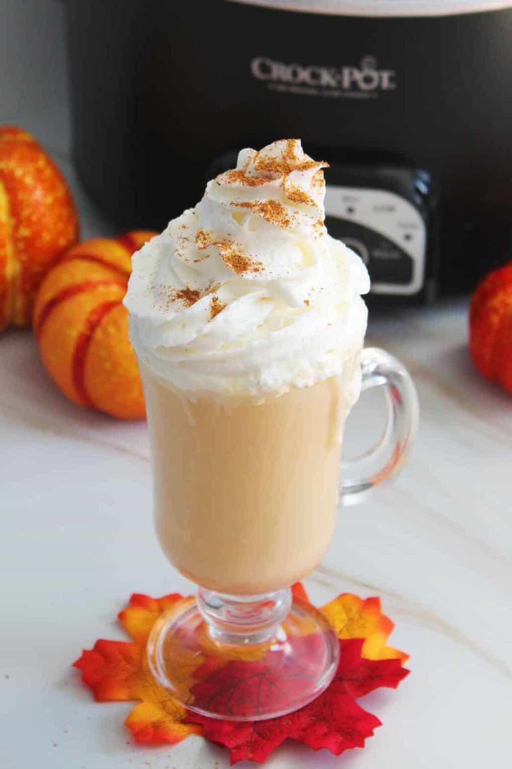 Light brown drink in a clear mug topped with whipped cream and some brown spice. Crockpot and mini pumpkins in the background.