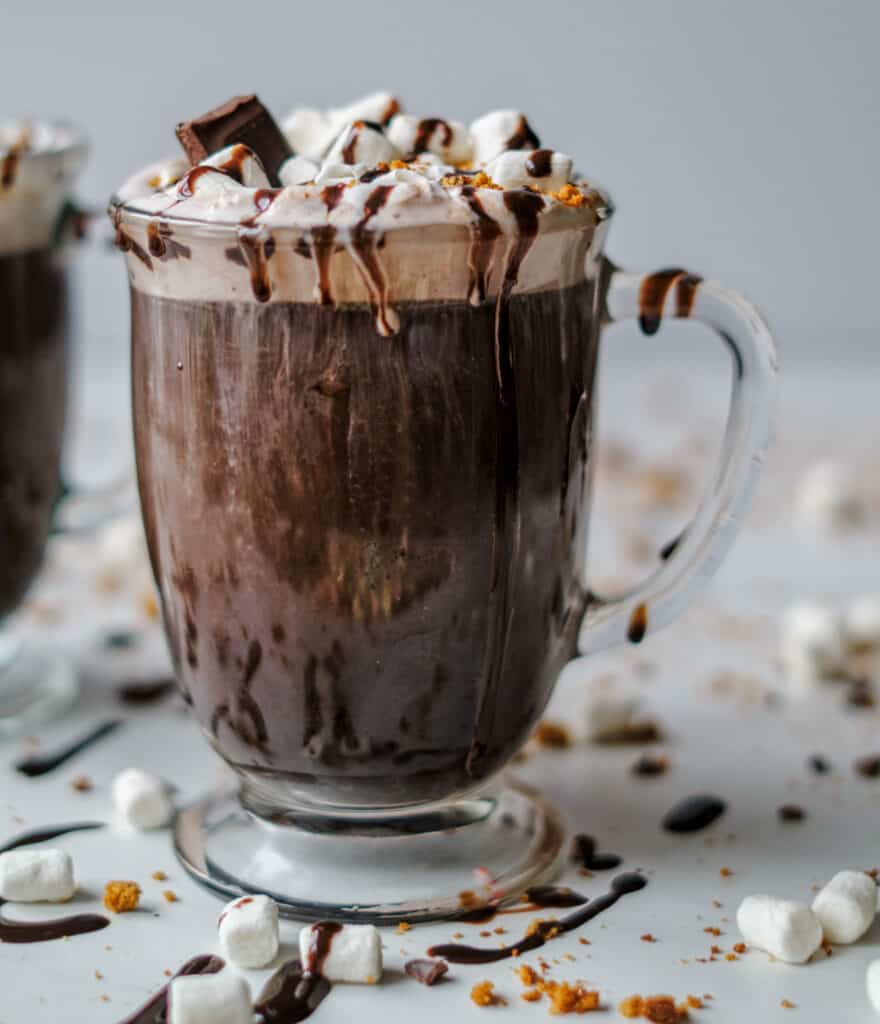 Clear mug full of hot chocolate. Topped with drizzled chocolate syrup, crushed graham crackers, and marshmallows.