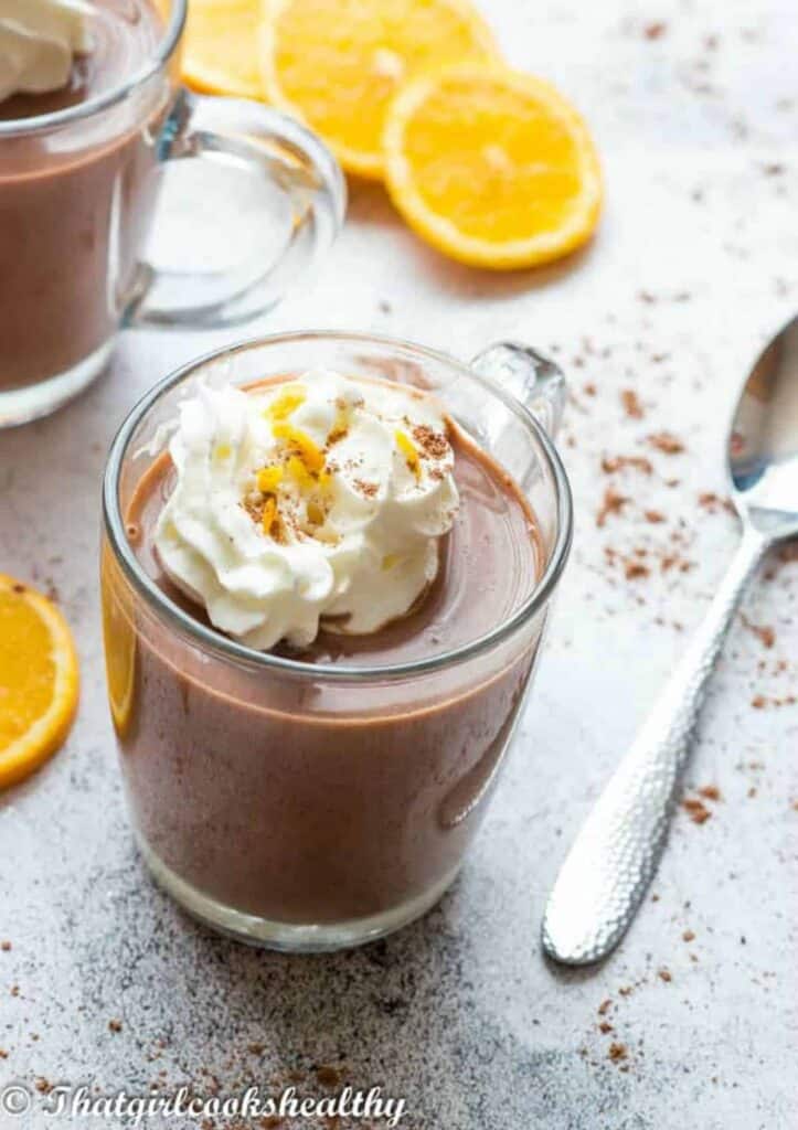 Clear mug of hot chocolate topped with whipped cream, sprinkled with cocoa powder and orange zest.