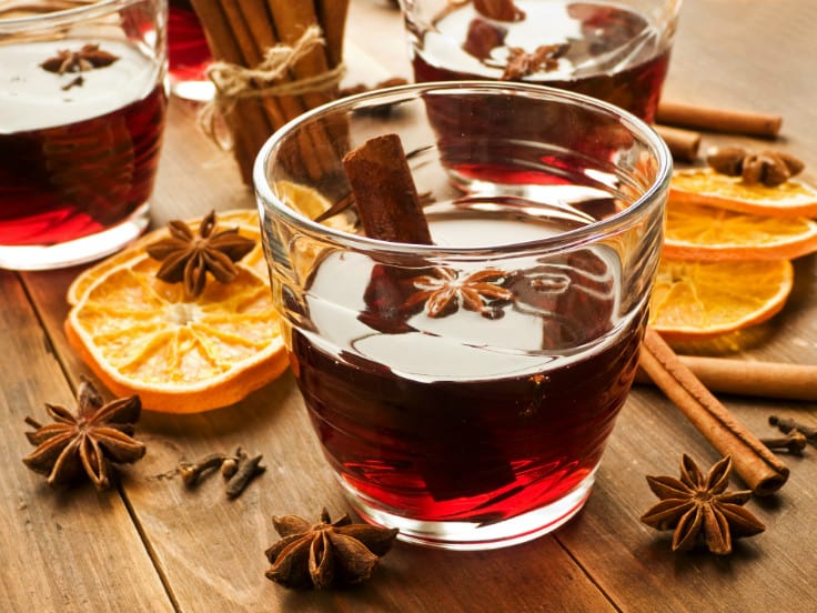 Deep red drink in clear mug with cinnamon stick. Sitting on a dark wood table with dried orange slices and spices scattered around.