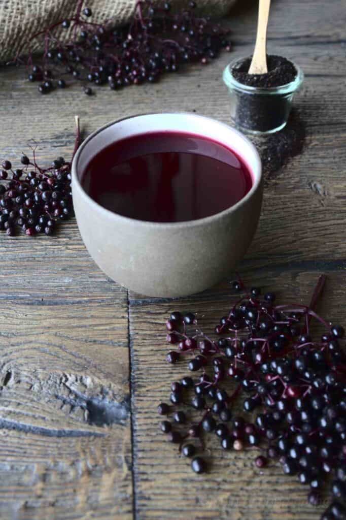 Clay mug of purple tea on a wood table with elderberries all around.