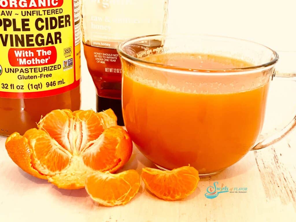 Orange drink in a clear mug sitting on a table with orange slices, a bottle of honey, and a bottle of apple cider vinegar.