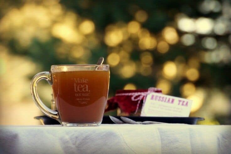 Clear mug of orange-brown tea sitting on a fabric covered table with blurred twinkle lights in the background.