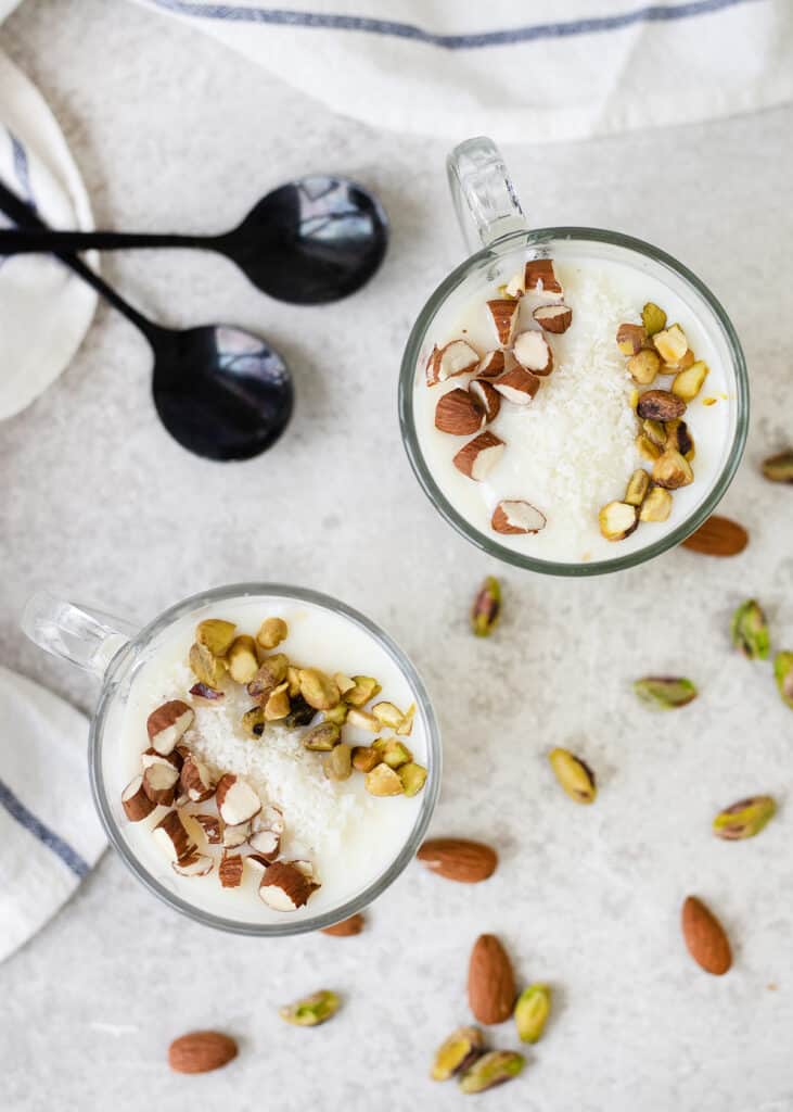 White drink in two clear mugs topped with chopped almonds, pistachios, and shredded coconut.