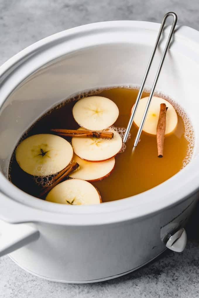 Apple cider being cooked in a white crockpot with apples slices and cinnamon sticks floating on top.