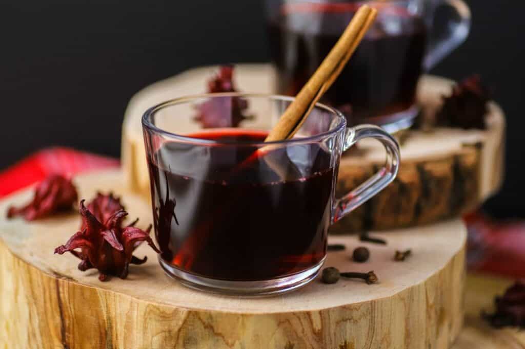 Clear mug of deep red drink with a cinnamon stick popping out. Sitting on a round cut of wood.