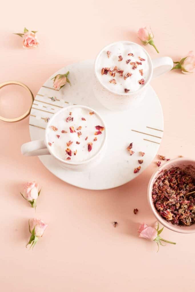 Two white, frothy drinks in white mugs, dried rose petals sprinkled on top. Sitting on a white tray on top of a pink table.