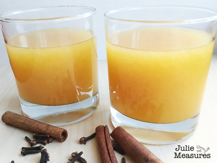 Two clear glasses full of orange drink.  Sitting on light wood with cinnamon sticks laying next to the glasses.