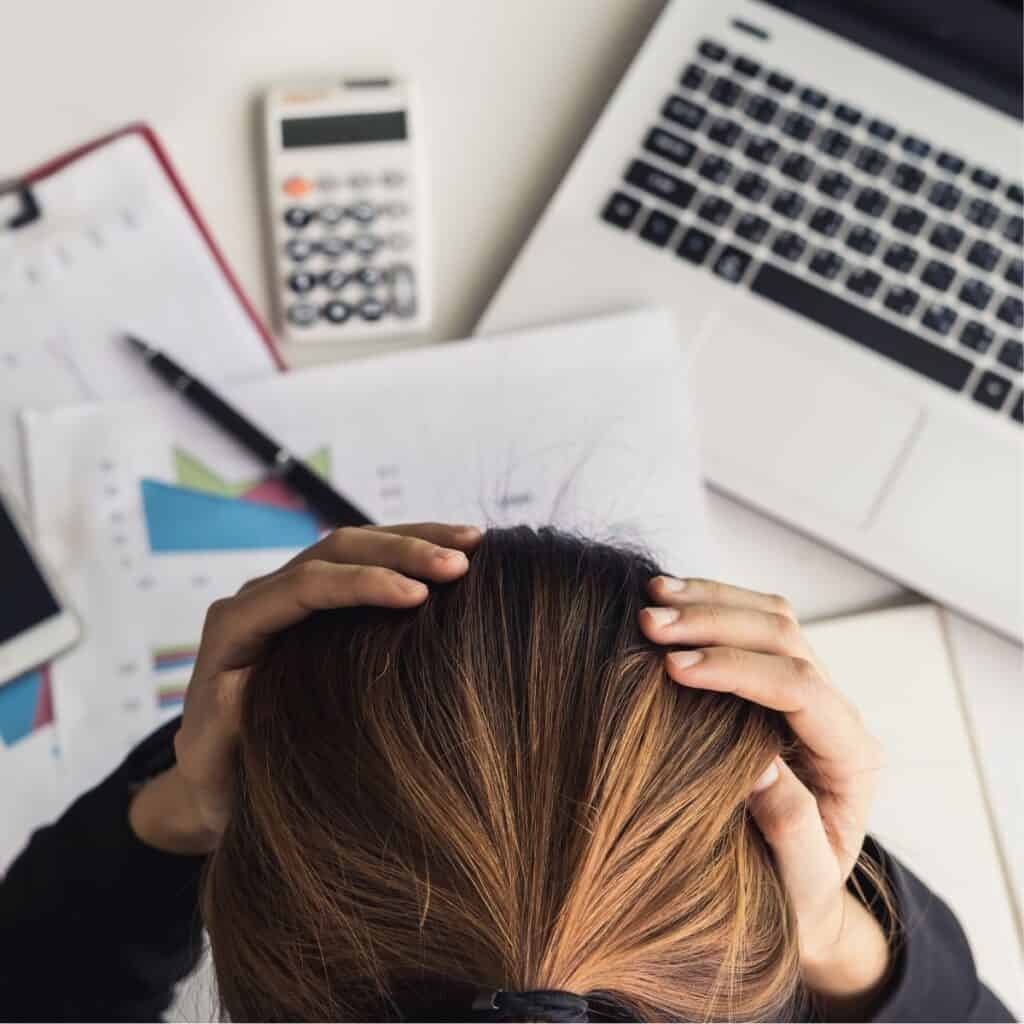 Woman sitting at a desk that's cluttered with paperwork, calculator, laptop. Her head is down, hands at her temples because she is stressed out.