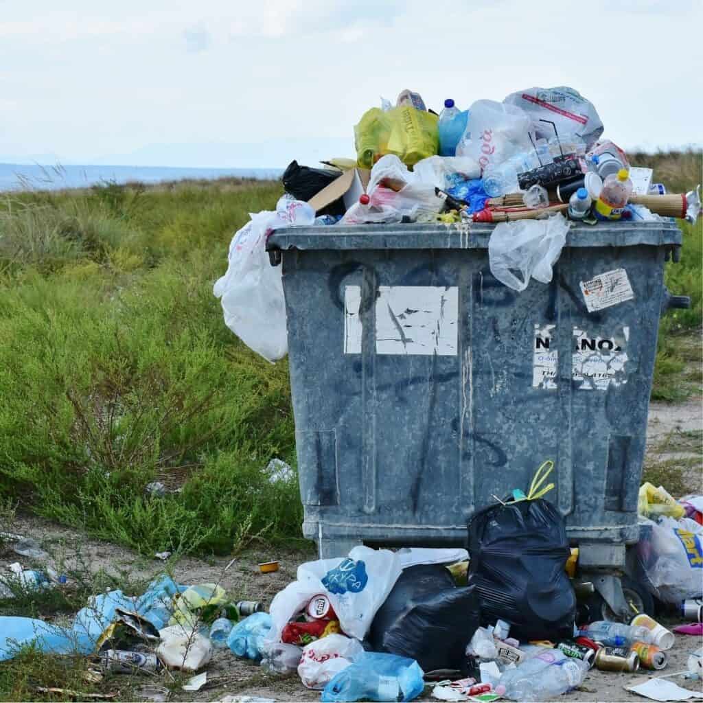 Overflowing trash bin in a grassy field with trash spilled out on the ground.