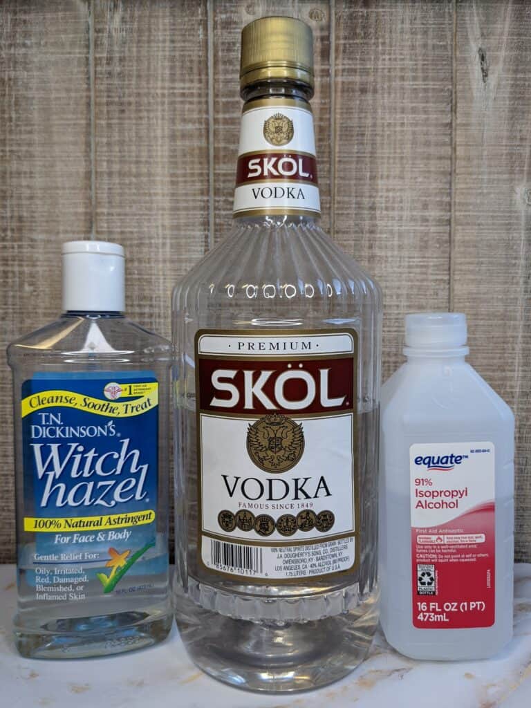 A bottle of witch hazel, vodka, and rubbing alcohol in front of a light brown, wood slat backdrop.