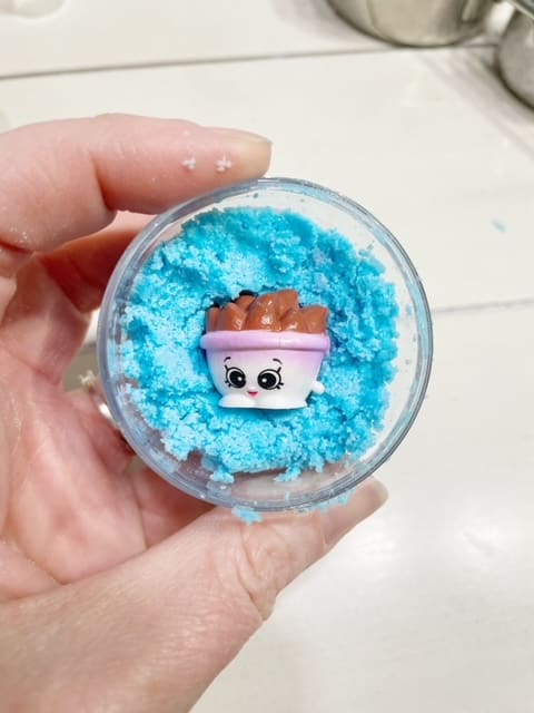 Picture of hand holding up half a bath bomb mold.  Their is blue bath bomb mixture inside and in the very center is a shopkins toy. | wethreeshanes