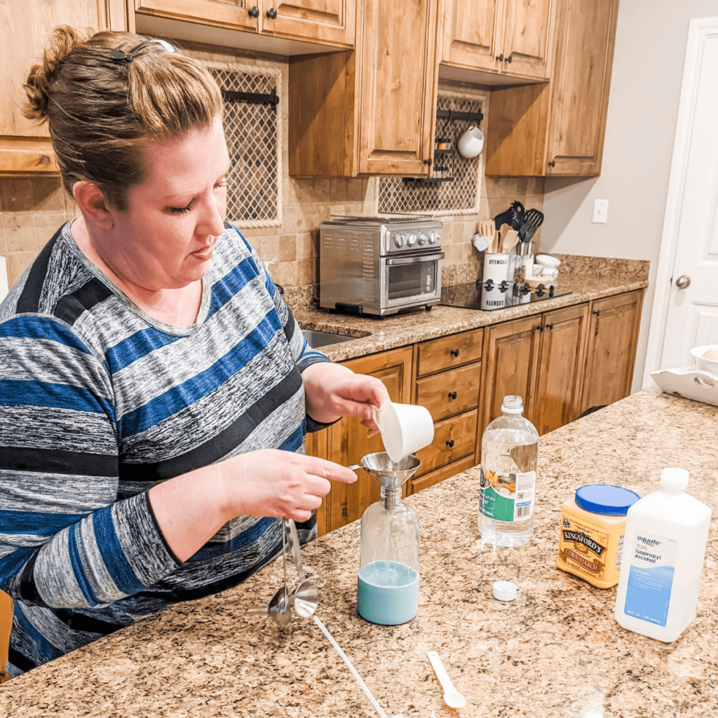 Caucasian woman in her 40's pouring a cup of clear liquid into a funnel that's propped in a glass bottle. She's making her own glass cleaner.