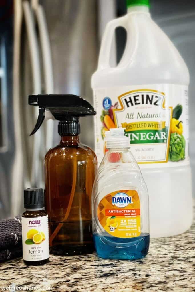 Homemade Stainless Steel Ingredients - White vinegar, dish soap, lemon essential oil, amber spray bottle and a grey cloth sitting on a counter top with a stainless steel fridge in the background.