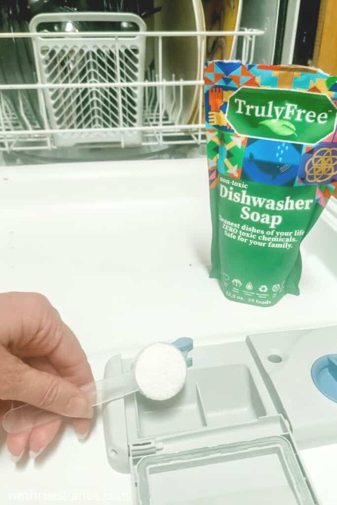 A full dishwasher with the door down. A package of Truly Free dishwasher soap sitting on the open door with a hand pouring a small scoop of powder soap into the dispenser.