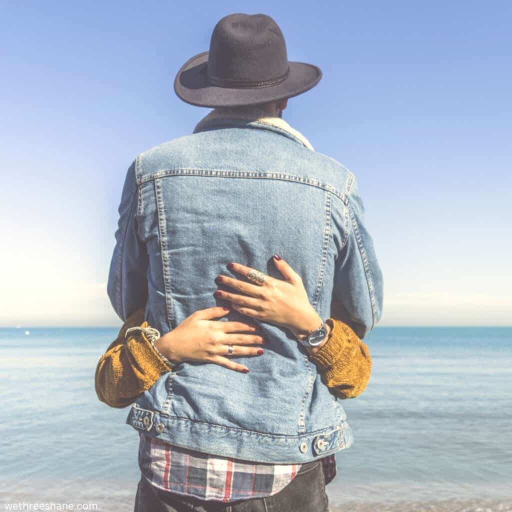 Back of a man standing on the beach, facing the water. Woman's arms wrapped around his waist in an imbrace.