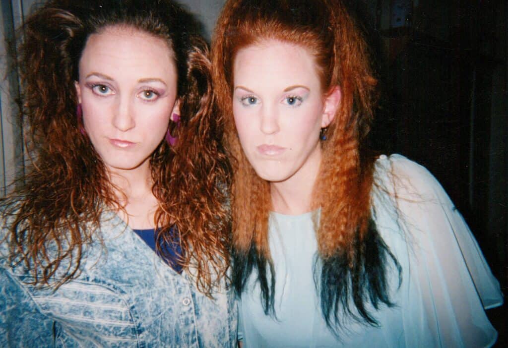 Two young adult women in 80s clothing with big hair and glossy pink lips.