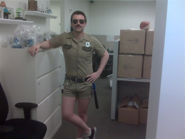 Guy dressed in beige button up, short sleeve shirt, tucked into short beige shorts. Wearing a big black belt with night stick and a big shiny badge on his chest.