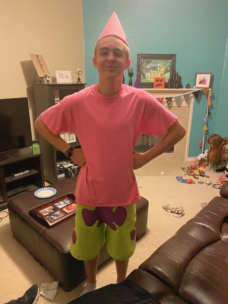 Teenage boy wearing a pink t-shirt, green shorts with purple blobs painted on them and a pick cone hat on his head to look like Patrick Star from Sponge Bob.