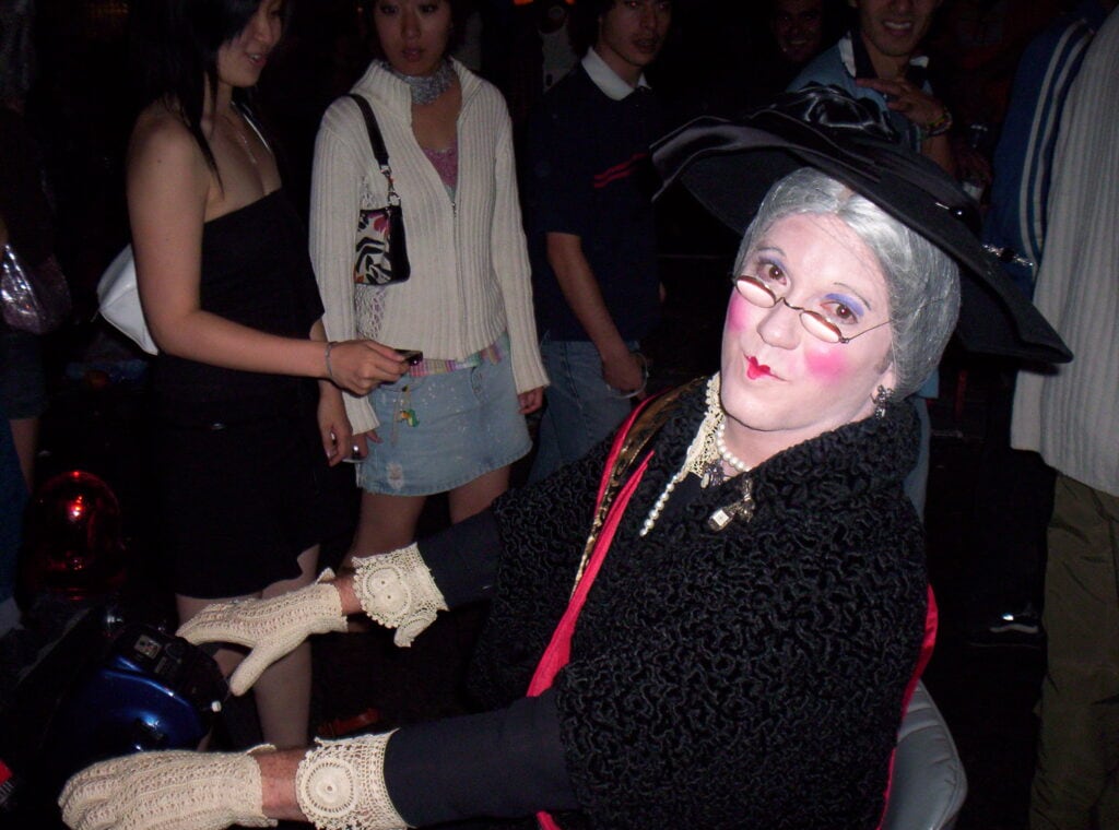 Guy dressed as an old lady riding a seated scooter in a Halloween parade.