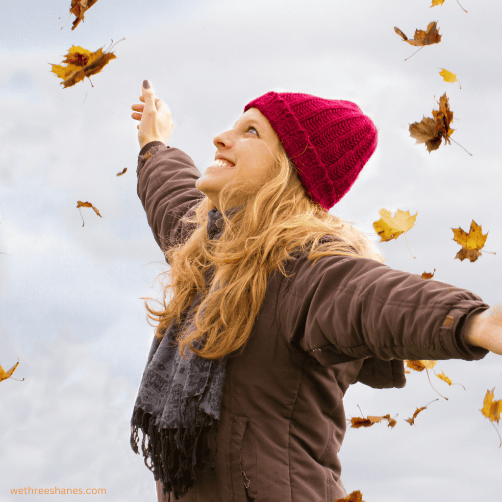 Lady dressed in a brown puff jacket, scarf and red knit beanie smiling in the falling leaves.