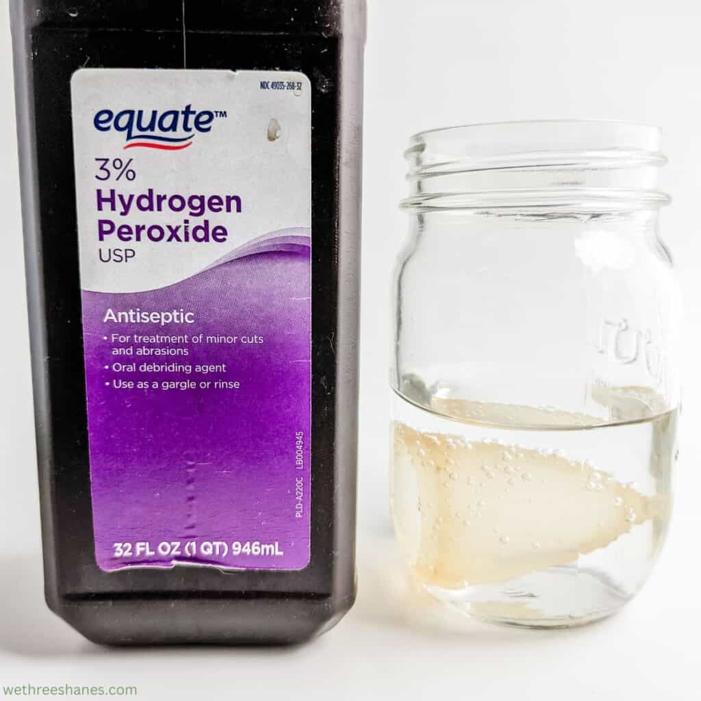 A bottle of hydrogen peroxide sitting next to a glass jar with a menstrual cup soaking in a solution of water and hydrogen peroxide.