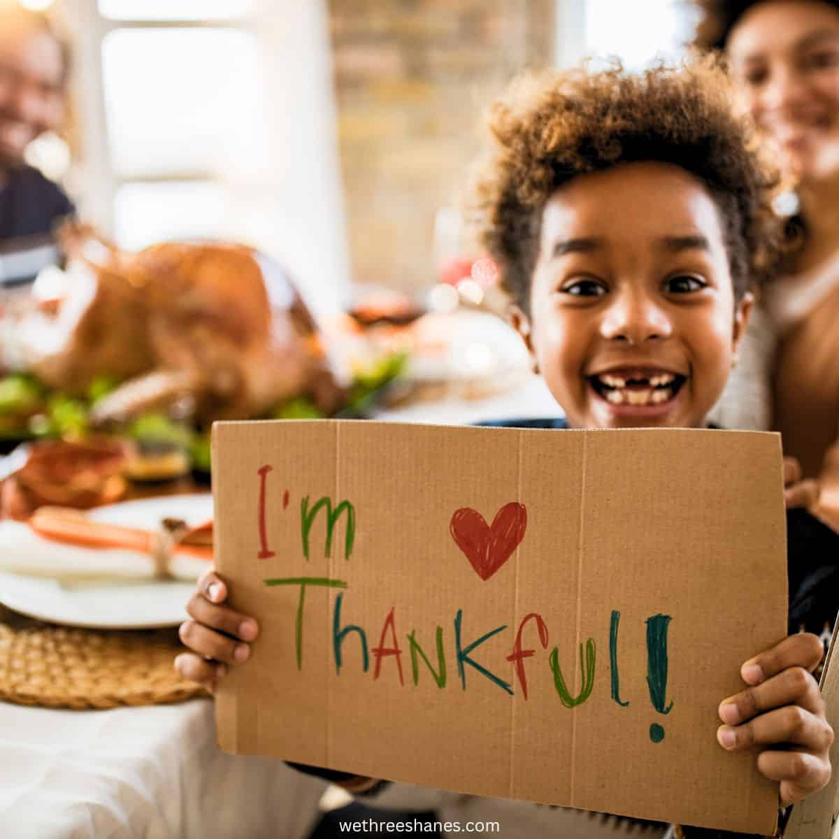 Young, smiling boy holding up a sign that reads, "I'm thankful!" with a thanksgiving dinner in the background.