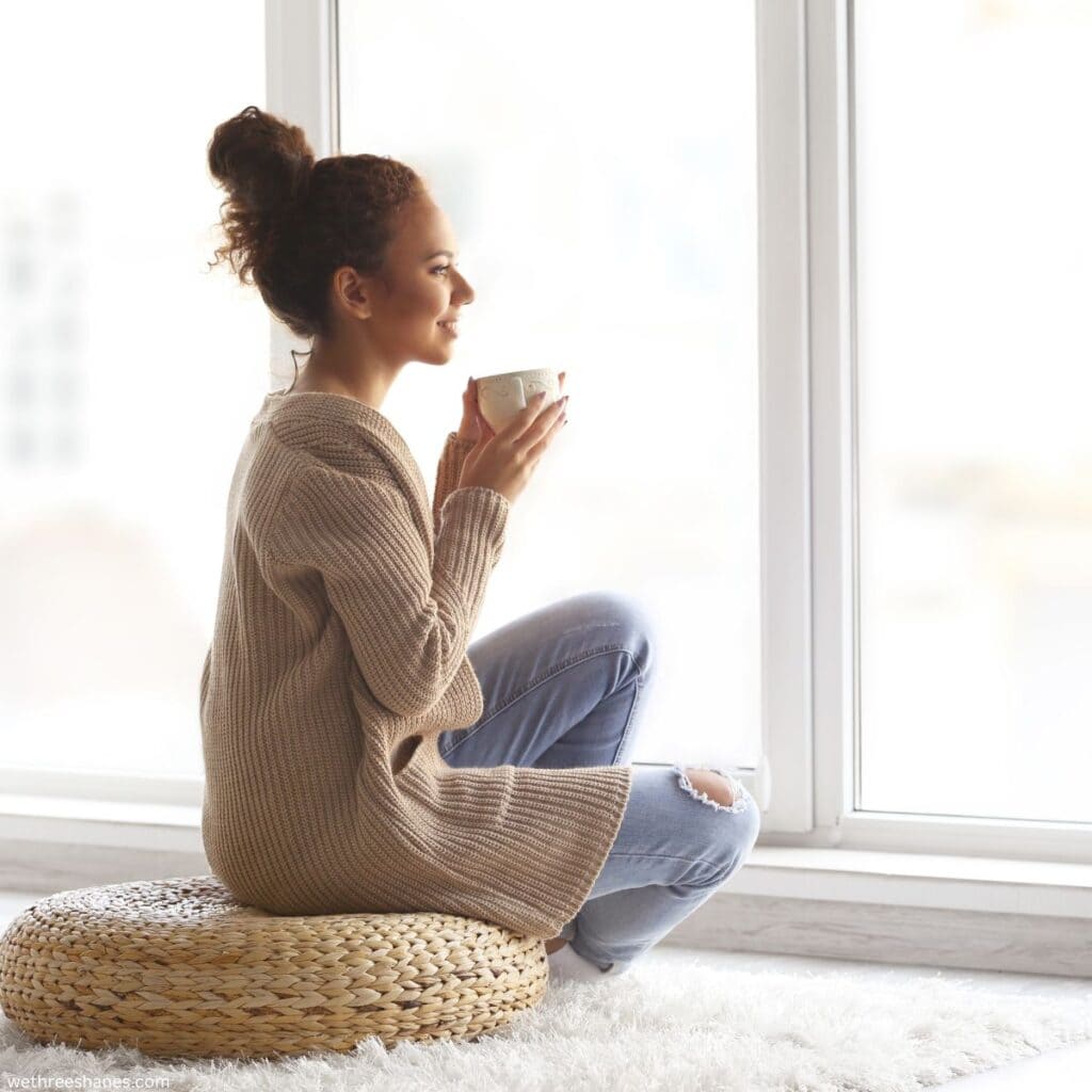Woman sitting on a mat, drinking from a mug, looking out the window